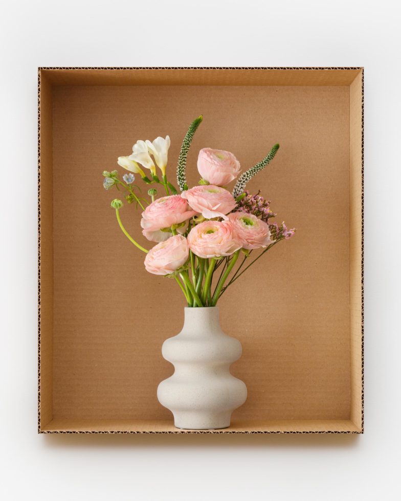 This is a photograph of a bouquet of flowers in a white vase inside a cardboard box.