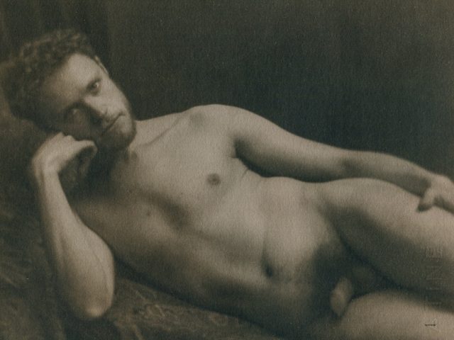 This is a sepia tone photograph of a nude man lying on his side holding his hand against his face staring at the camera.