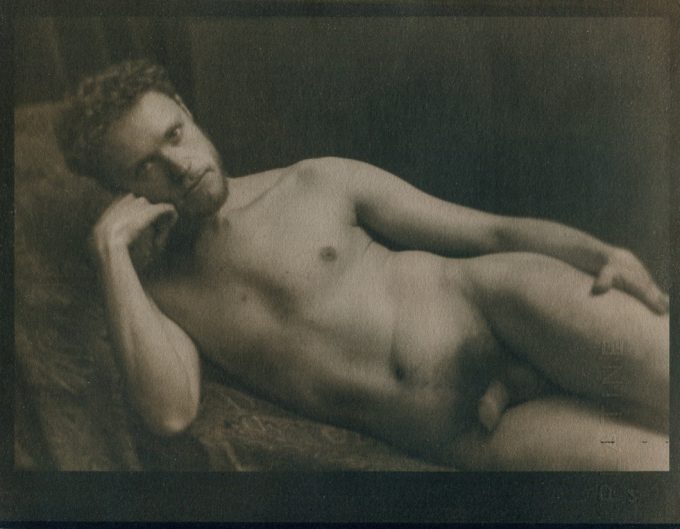 This is a sepia tone photograph of a nude man lying on his side holding his hand against his face staring at the camera.