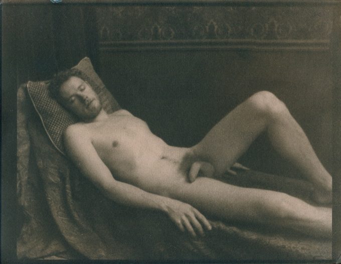 This is a sepia tone photograph of a nude man lying on a chaise lounge with his left knee bent.