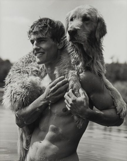 This is a black-and-white photograph of a smiling shirtless man with a golden retriever on his shoulders.