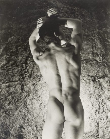 This is a black-and-white photograph of a nude man from behind with his arms raised above his head in what appears to be a cave of some kind.