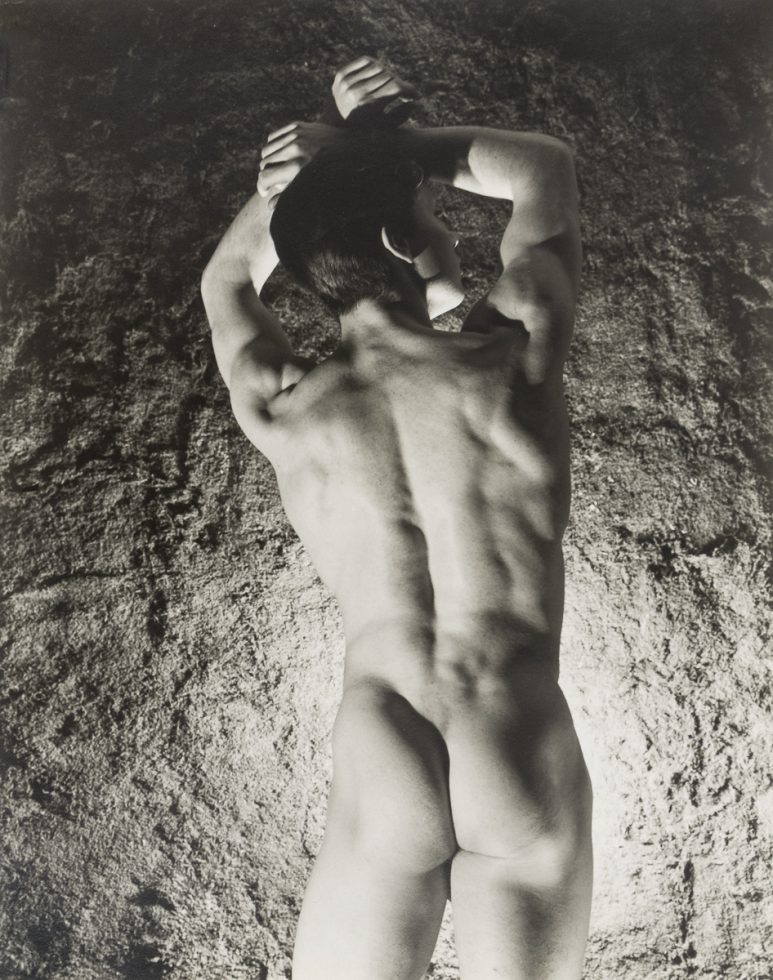 This is a black-and-white photograph of a nude man from behind with his arms raised above his head in what appears to be a cave of some kind.