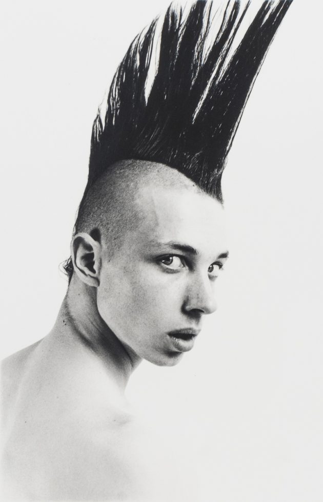 A black-and-white photograph of a boy with a large black mohawk looking back over his right shoulder.