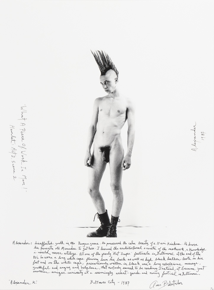 This is a black and white photograph of a naked young man with a mohawk and black boots standing in a white studio setting.
