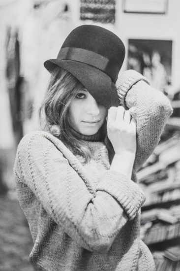 This is a black-and-white photograph of a woman in a sweater holding a black hat over her left eye.