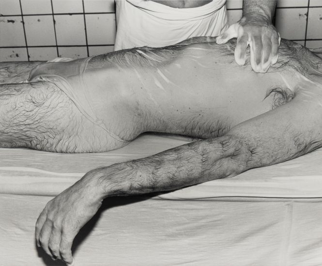 This is a black and white photograph of a hairy and wet man receiving a massage in a bathhouse.