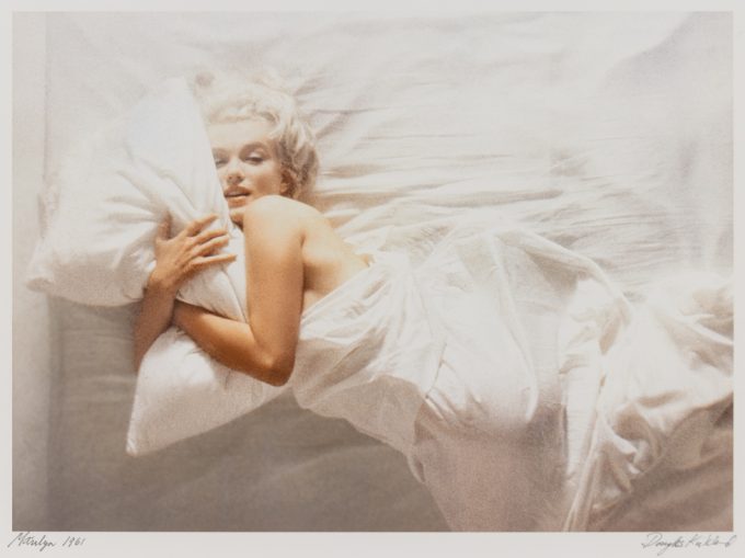 This is a color photograph of a platinum blonde woman in a bed with white sheets.