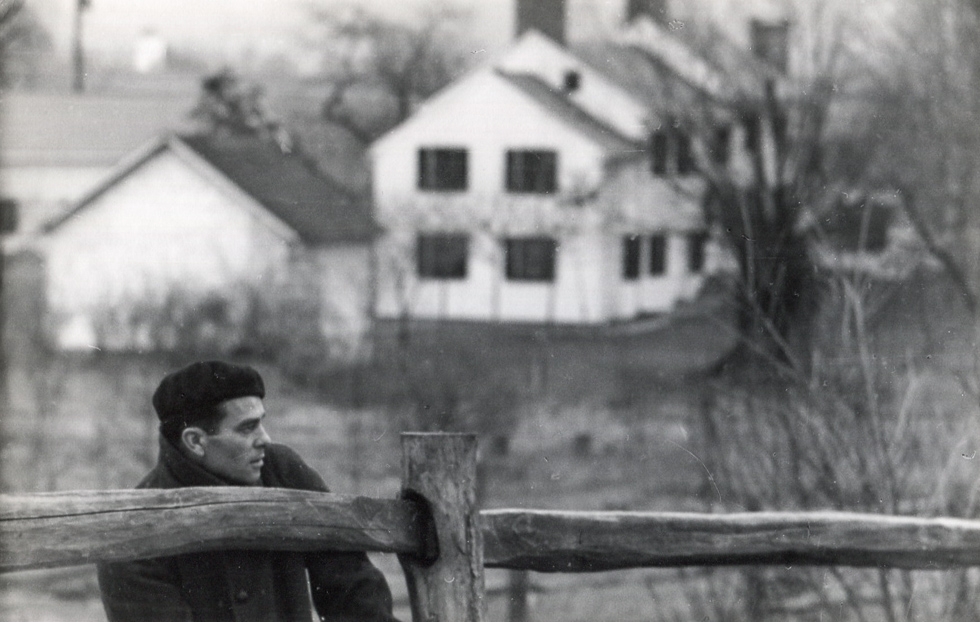 This is a black and white photograph of a handsome man with dark hair by a wooden fence in a country setting.