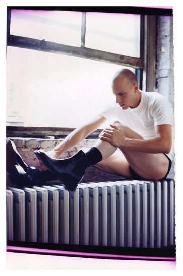 This is a photograph of a man in leather booths, underwear, and a white tee shirt sitting on a radiator next to a window.