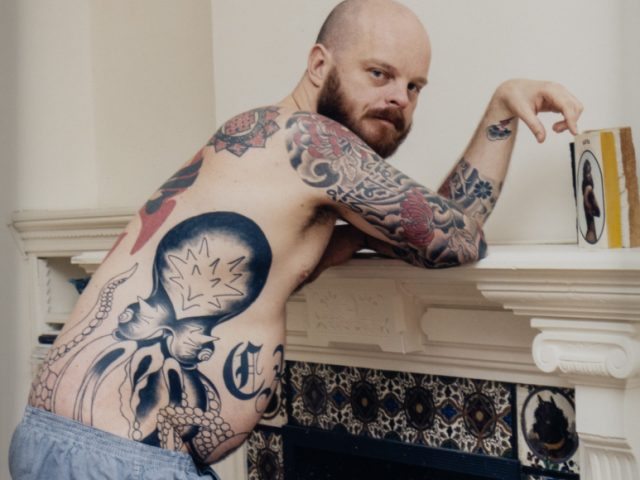 This is a color photograph of a Male with Tattoos Leaning Against a Fireplace.