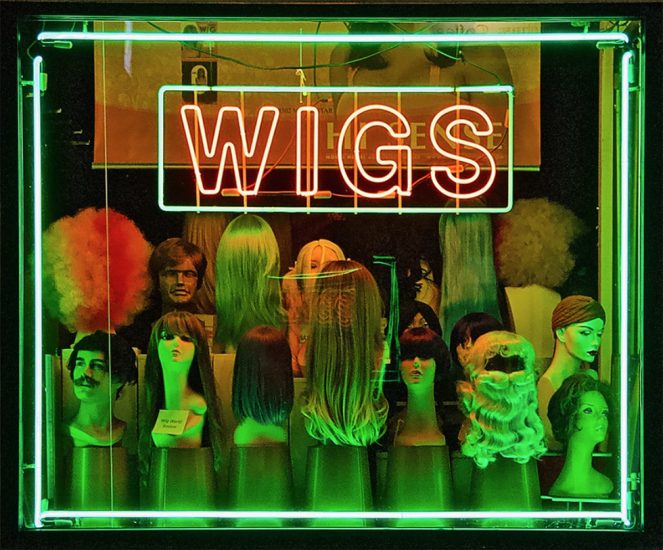 This is a photograph of wigs on display in a green neon-lit window with a red neon sign that reads "WIGS"