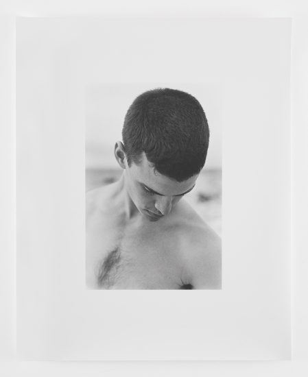 This is a photograph of a young man with short dark hair on a beach with his head looking down towards the ground.