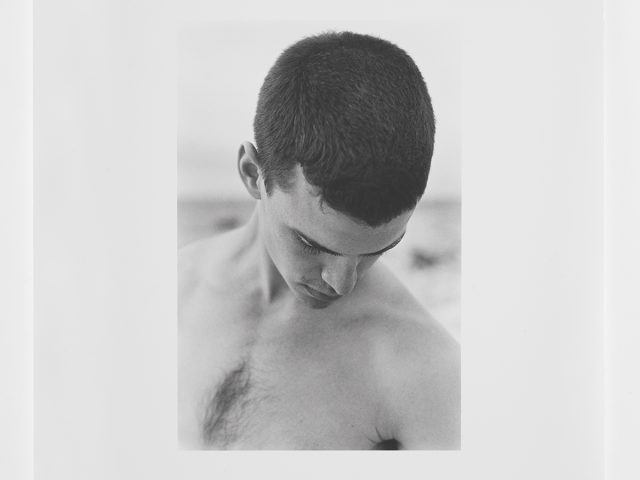 This is a photograph of a young man with short dark hair on a beach with his head looking down towards the ground.