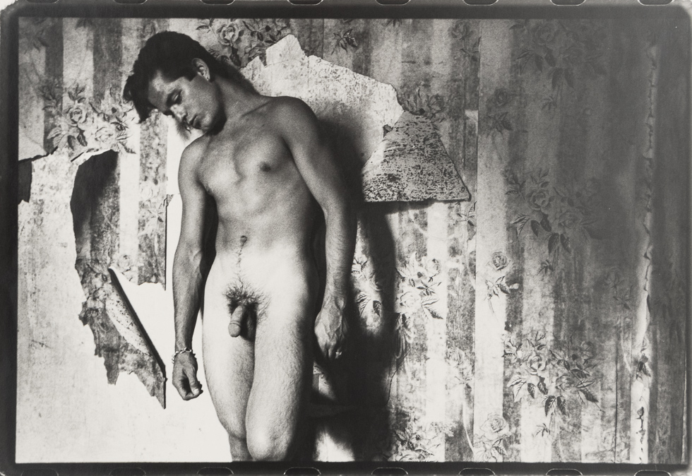 This is a black and white photograph of a nude man standing beside a distressed wall.