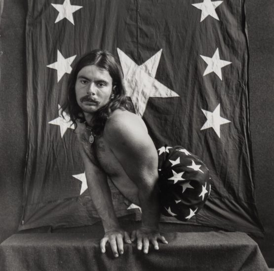 This is a photograph of a man squatting wearing no shirt and pants with stars on them against a starred backdrop.