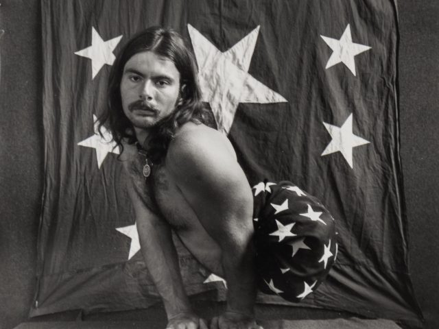 This is a photograph of a man squatting wearing no shirt and pants with stars on them against a starred backdrop.