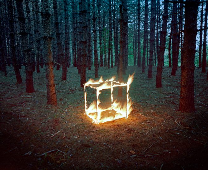 This is a photograph of a cube on fire in the woods at dusk.