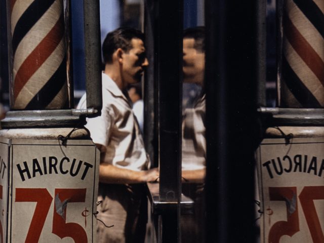 This is a color photograph of a man looking in the glass facade of a barber shop.