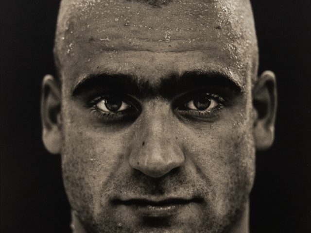 This is a photograph of a man with dark, thick eyebrows, staring straight into the camera in front of a black backdrop.