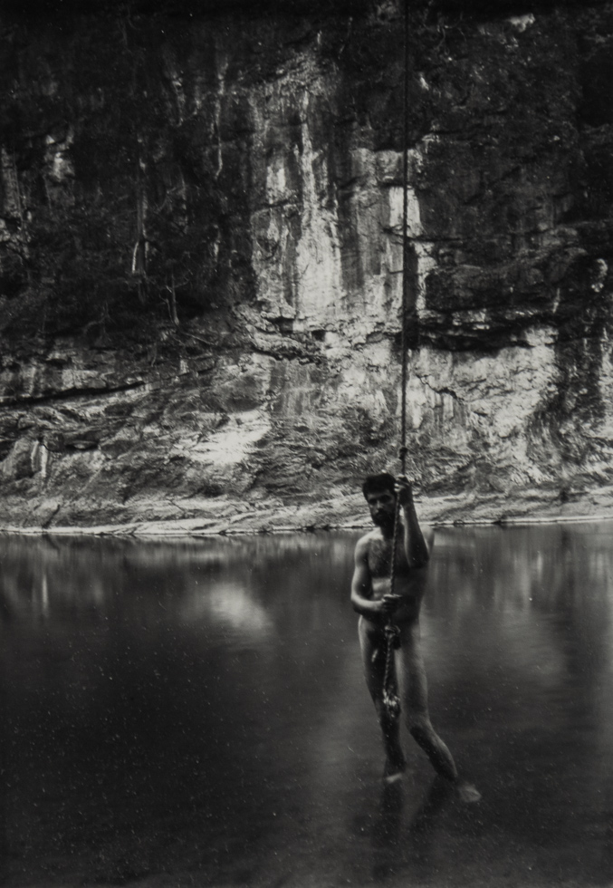 This is a black and white photograph of a nude man standing in a body of water in front of a wall of rock and greenery.