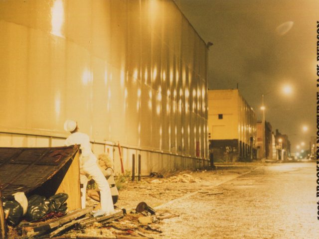 This is a photograph of a man receiving oral sex in an industrial area, wearing a navy uniform.