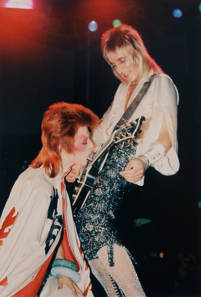 This is a photograph of two musicians performing on stage, with one playing a guitar with their teeth.