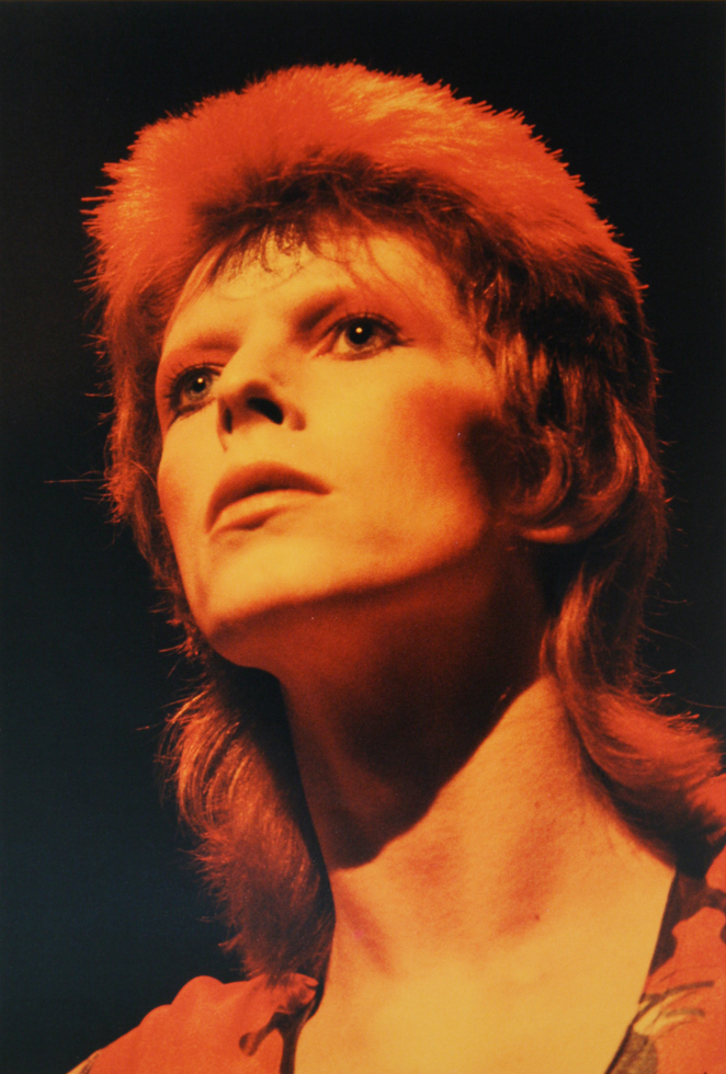 This is a photograph of a David Bowie bathed in golden light against a black background.