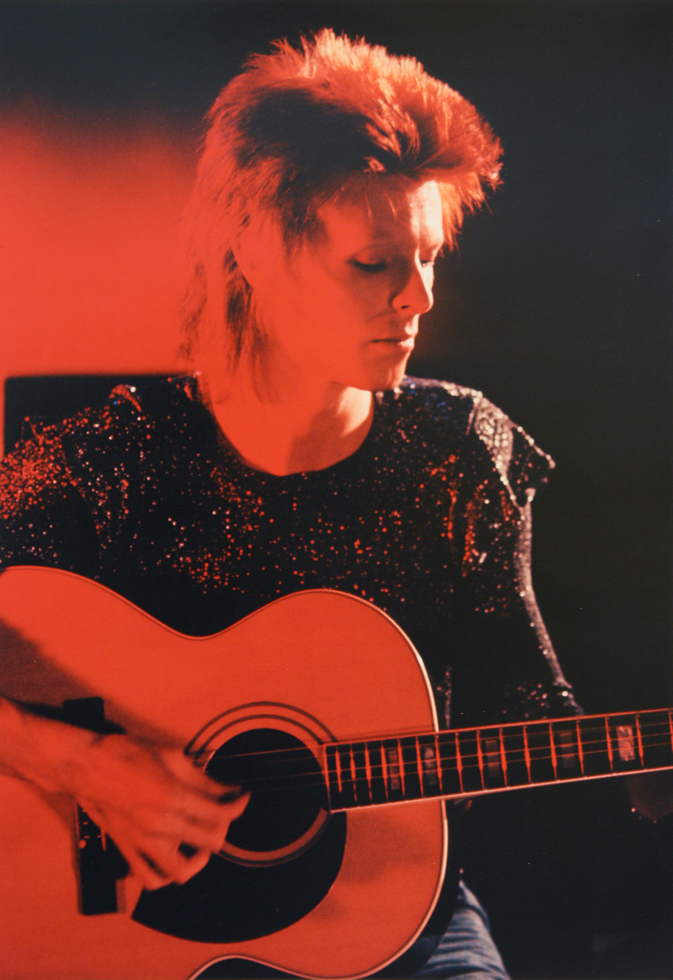 This is a black and red photograph of David Bowie playing an acoustic guitar.