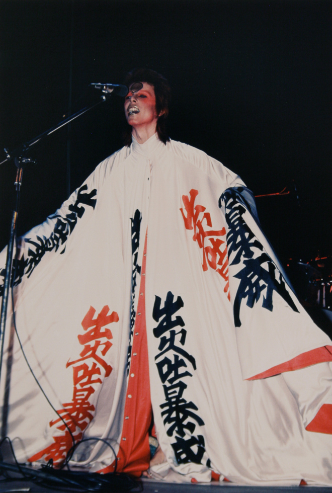 This is a photograph of David Bowie wearing a white kimono standing in front of a microphone