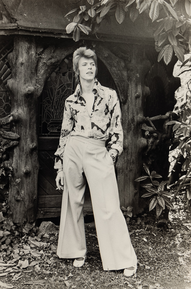 This is a black and white photograph of David Bowie wearing flaired pants.