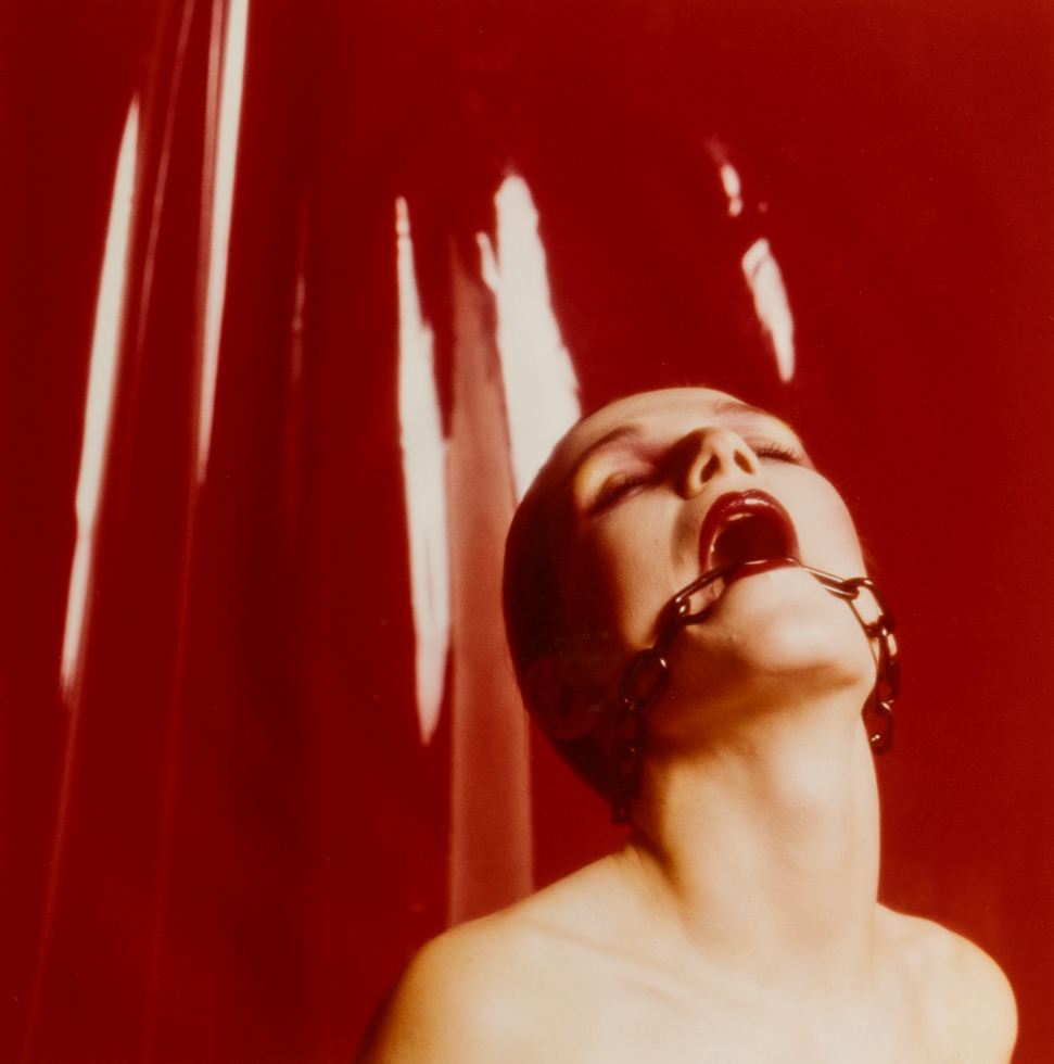 This is a color photograph of a person with a gag in their mouth against a red backdrop.