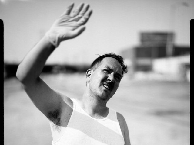 This is a photograph of a man in a white tank top wearing a latex glove, shielding his eyes from the sun with an outstretched arm.