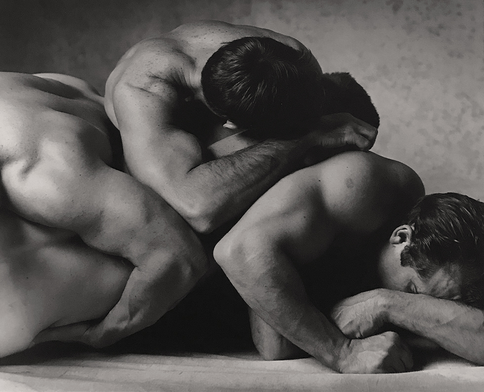 This is a black and white photograph of nude men embracing with three elbows touching one another.