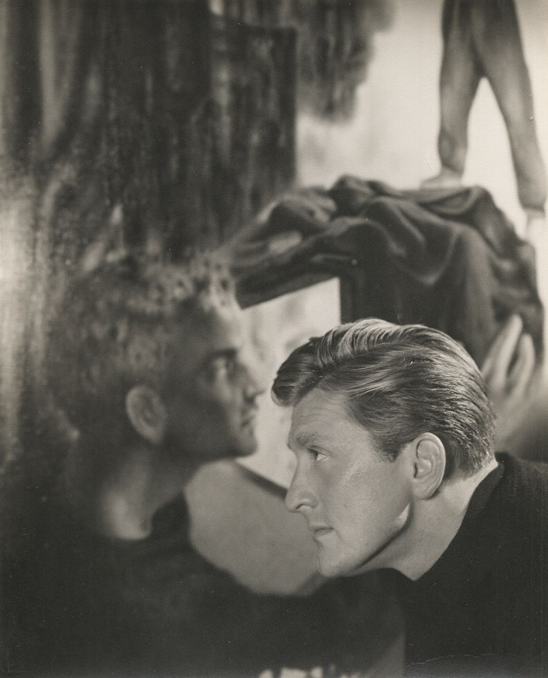 This is a black-and-white photograph of a handsome man in profile against a setting of a portrait bust and a painting.