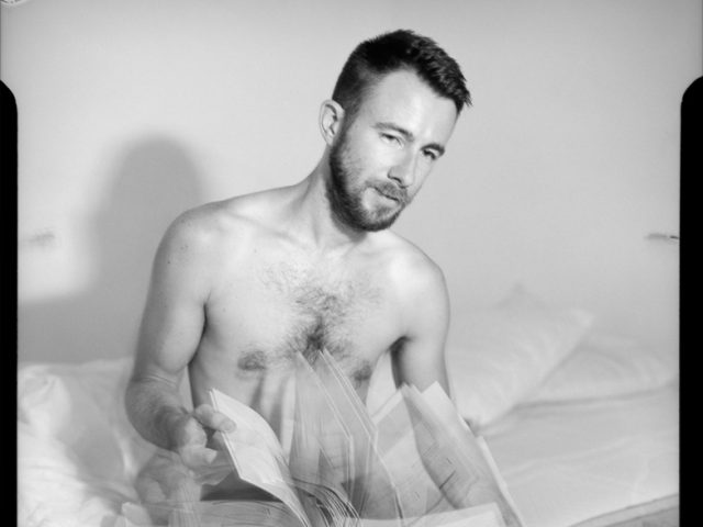 This is a black and white photograph of a shirtless man flipping through a coffee table book.