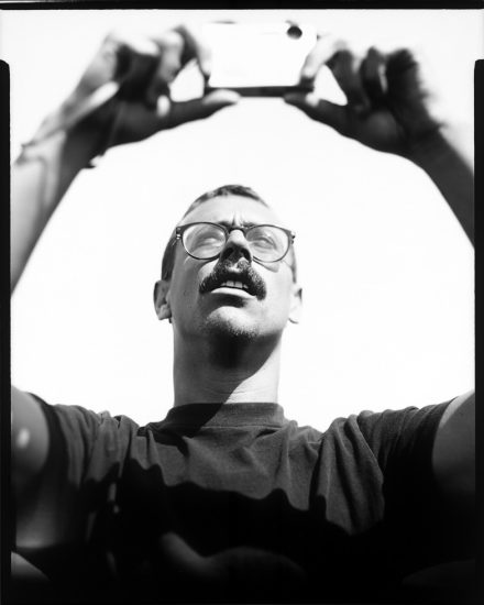 This is a black and white photograph of a man taking a picture on a point and shoot camera with a bright white sky behind him.