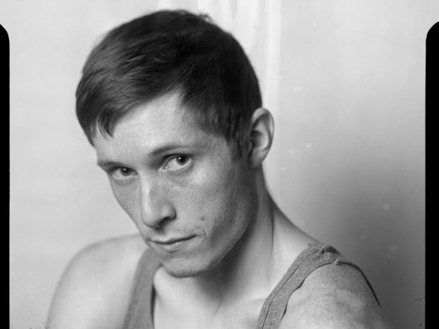 This is a photograph of a young man in a tank top looking directly at the camera.