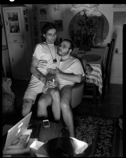 This is a black and white photograph of two men in white shirts sitting on a chair in a living room illuminated by a camera flash.