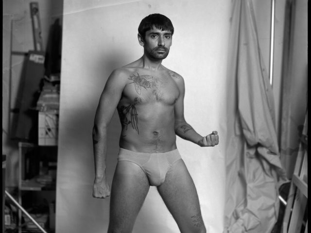 This is a black and white photograph of a shirtless man in front of a white backdrop in a photo studio.