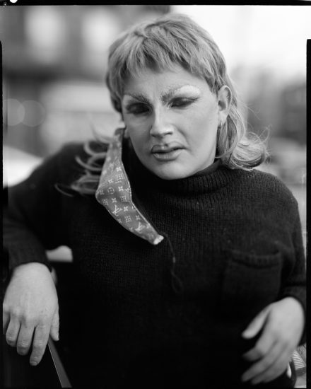 This is a black and white photograph of a person in drag makeup with a face mask dangling from their ear.