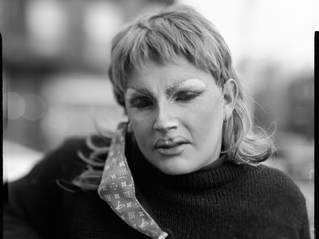 This is a black and white photograph of a person in drag makeup with a face mask dangling from their ear.