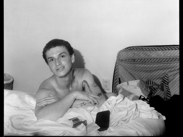 This is a black and white photograph of a young man next to a wall with posters.