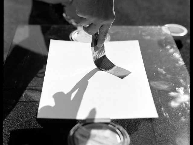 This is a black and white photograph of a piece of paper with a hand brushing on photosensitive chemicals.