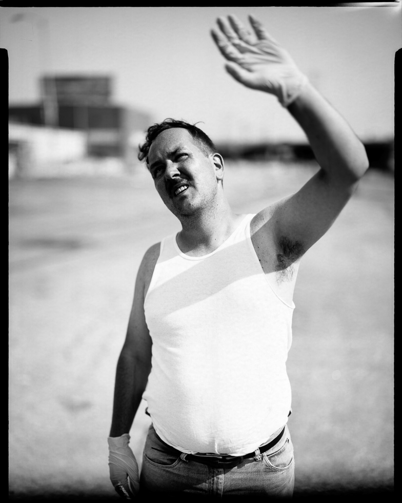 This is a photograph of a man in a white tank top wearing a latex glove, shielding his eyes from the sun with an outstretched arm.