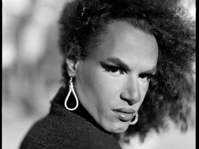 This is a black and white photograph of a person in a park wearing a black jacket with long silver earrings looking at the camera with an intense expression.