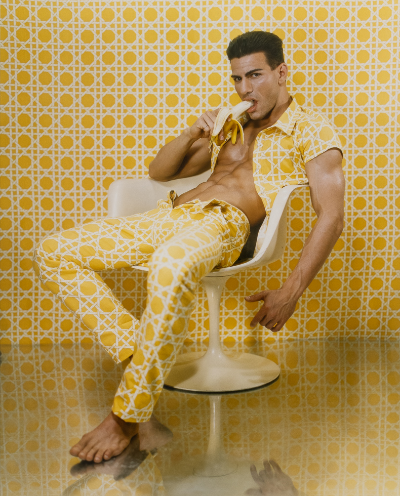 This is a color photograph of a handsome man sitting in a tulip chair eating a banana.