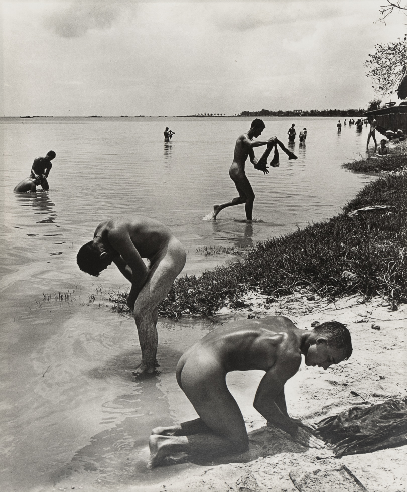 This is a black-and-white photograph of men nude on a beach and in the water.