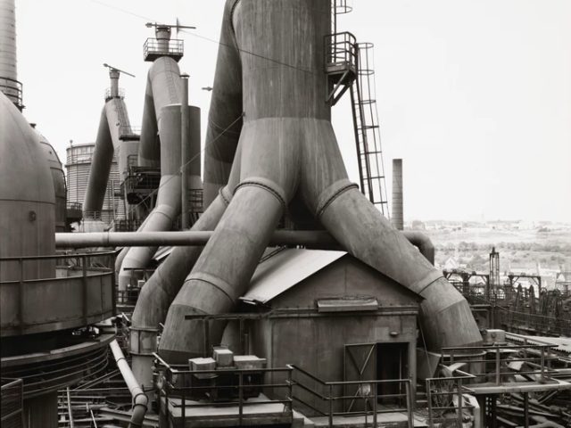 This is a black-and-white photo of an industrial steel mill with towering blast furnaces and complex piping.