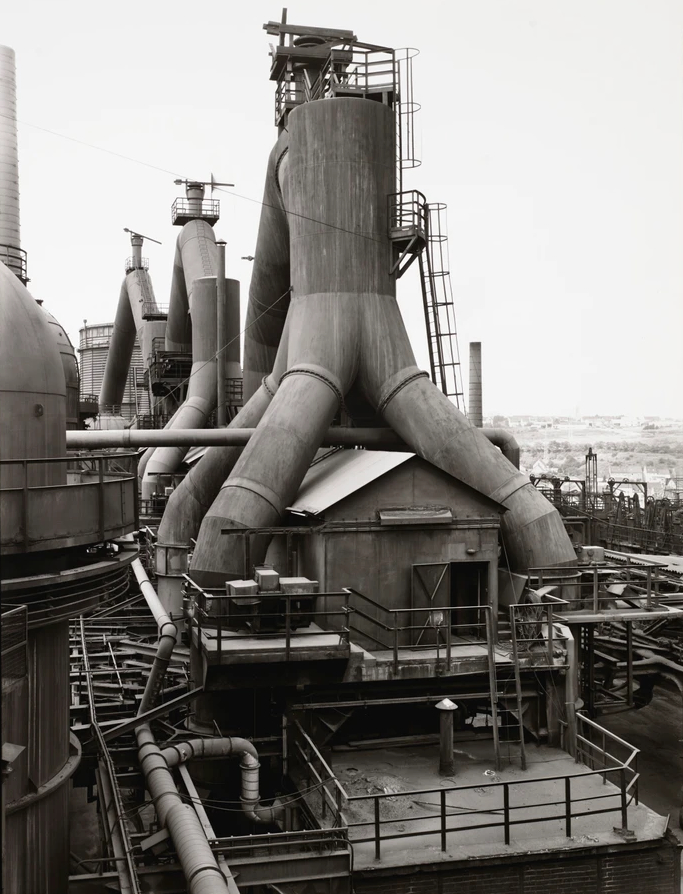 This is a black-and-white photo of an industrial steel mill with towering blast furnaces and complex piping.